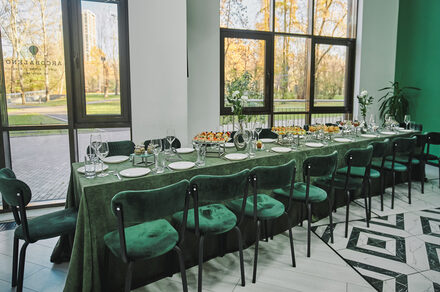 Arcobaleno Loft(Аркобалено Лофт). Пространство с длинным столом, покрытым зеленой скатертью, окружено стульями с зелеными бархатными сиденьями. На столе стеклянные бокалы, белые тарелки и закуски. Большие окна с видом на парк. Пол выложен узорчатой плиткой.