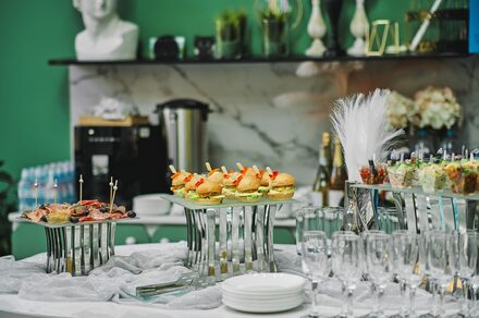 Arcobaleno Loft(Аркобалено Лофт). Праздничное пространство со столом для фуршета. На столе бутерброды, закуски в стеклянных стаканах, десерты, стеклянные бокалы. На заднем плане декор и напитки, зеленые стены, бело-мраморная столешница.