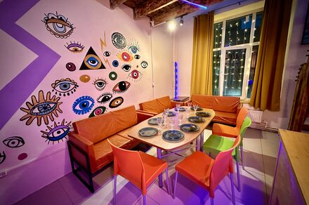 Яркий двухуровневый лофт. Стол с посудой. Яркие стулья с диванами. Обклеенные рисунками стена. Окно с навешенной гирляндой