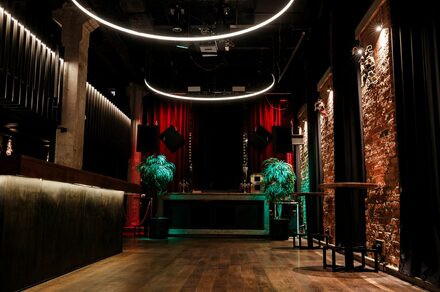Treff8 Club (Трефф8 Клуб). Двухуровневый клуб под закрытие. Пространство с деревянным полом, барной стойкой и кирпичными стенами. Красные занавесы на сцене и декоративные растения. Подвесные кольцевые светильники и точечное освещение. Черные элементы декора, металлические колонны и подвесные динамики.