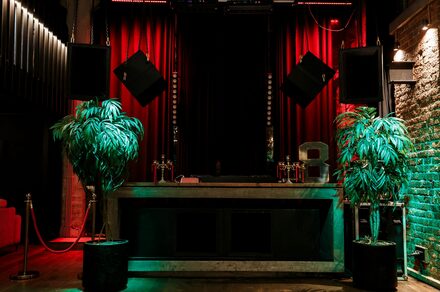 Treff8 Club (Трефф8 Клуб). Двухуровневый клуб под закрытие. Темное праздничное пространство с барной стойкой из бетона, двумя крупными колонками по бокам, богатой драпировкой из красных занавесок. По обе стороны от стойки стоят высокие растения в горшках. Стена из кирпича, освещенная красным и зеленым светом.