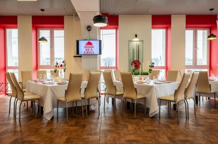 Sea Grill(Морской гриль). Лофт-бар под закрытие с видом на Неву. Банкетный зал с двумя круглыми столами, накрытыми белыми скатертями, окруженными бежевыми стульями. Зона с темно-коричневым деревянным полом, бело-красными стенами и окнами. Панельный потолок с черными светильниками. На стене телевизор с логотипом «SEA GRILL».