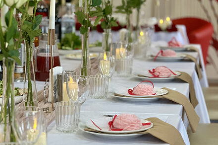 Sea Grill(Морской гриль). Лофт-бар под закрытие с видом на Неву. Длинный стол с белой скатертью, сервированный белыми тарелками с розовыми декорациями и прозрачными бокалами. На столе зажжённые свечи в стеклянных подсвечниках и вазы с зелёными растениями. На заднем плане красные стулья, мягкое освещение.