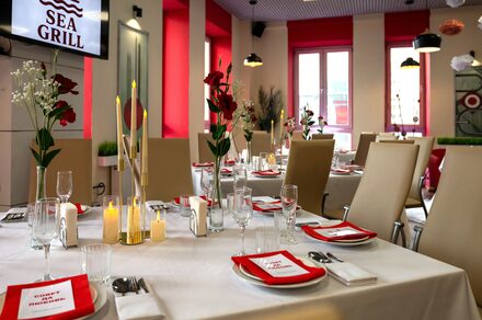 Sea Grill(Морской гриль). Лофт-бар под закрытие с видом на Неву. Столовая с накрытым белой скатертью длинным столом, украшенным красными салфетками и цветами в вазах. Кремовые кожаные стулья, на стене логотип «SEA GRILL», висят светильники. Яркие акценты красного цвета в интерьере.