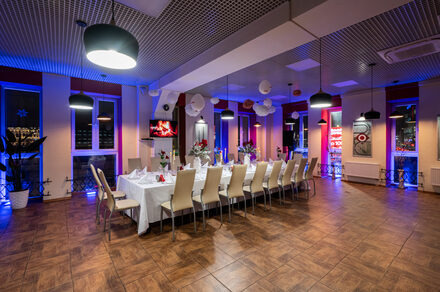 Sea Grill(Морской гриль). Лофт-бар под закрытие с видом на Неву. Просторная комната с длинным прямоугольным столом, накрытым белой скатертью, окруженным кремовыми стульями. Потолок с сетчатой структурой и подвесными светильниками. Пол плиточный, коричневый. На стенах окна, большие красные неоновые знаки.