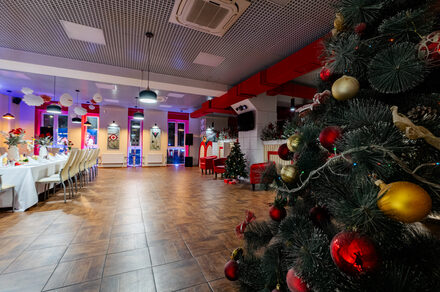 Sea Grill(Морской гриль). Лофт-бар под закрытие с видом на Неву. Большое помещение с елкой на переднем плане, украшенной красными и золотыми шарами. Справа столы с белыми скатертями и стульями. Потолок с подвесными светильниками и красными элементами. Паркетный пол, яркое освещение.