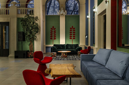 Viola Loft (Виола Лофт). Просторный и светлый лофт с панорамным видом в парк. Просторное помещение с высокими потолками, большими арочными окнами и зелеными акцентными стенами. Мягкая мебель: синие и зеленые диваны, красные кресла. Деревянный стол, декоративные элементы. Люстры и настенные светильники. Напольное покрытие из светлого материала.