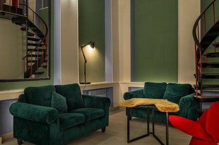Viola Loft (Виола Лофт). Просторный и светлый лофт с панорамным видом в парк. Двухуровневое помещение с винтовой лестницей, двумя тёмно-зелёными диванами и деревянным столом. Освещение обеспечивается настенным светильником. Стены окрашены в зелёный и белый цвета. На полу серые плитки.