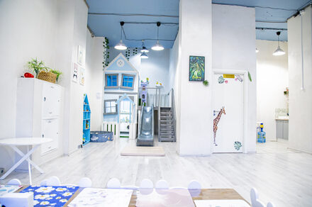 Лофт Облака. Зал Баунти на Полярной. Пространство детской игровой с зоной для занятий и горкой, большая белая комната с высоким потолком, белые стены и пол. Голубая игровая структура в виде домика с лестницей и горкой. Яркие акценты: жираф на двери, растения в горшках. Освещение подвесное.