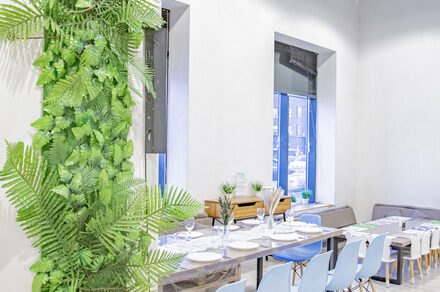 Лофт Облака. Зал Баунти на Полярной. Просторное помещение с белыми стенами, большое окно, стол с синей и серой мебелью. На столе сервировка с тарелками и бокалами. Вертикальная стена с зелеными растениями. Пол светлый. Дневное освещение.