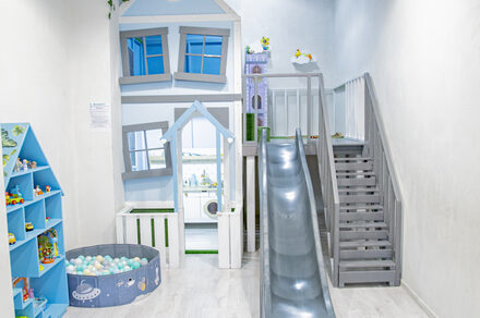 Лофт Облака. Зал Баунти на Полярной. Детская игровая зона с деревянным домиком, лестницей и горкой. Стены и пол светлые, корзина с цветными шарами. Шкаф с игрушками слева, домик окрашен в голубые и белые тона. Яркое освещение, на стене заметен текст.