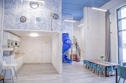 Лофт Облака. Зал Баунти на Сельскохозяйственной. Открытое пространство с высокими потолками, игровой горкой, зоной кухни и длинным столом с мягкими бирюзовыми стульями. Стены белые с синим узором, полы светлые. Естественное и искусственное освещение, большие окна, деревянная лестница.