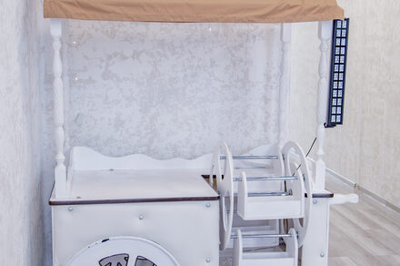 Лофт Облака. Зал Баунти на Сельскохозяйственной. Деревянная тележка с белыми и светло-коричневыми элементами, расположена у светлой стены с текстурой. Рядом светлая поверхность пола с деревянным узором. На тележке есть полки и стилизованные колеса.