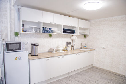 Лофт Облака. Зал Баунти на Сельскохозяйственной. Кухонное помещение с белыми шкафами и светлыми столешницами. На столе микроволновка, термопот и кухонные принадлежности. Открытые полки с посудой. Белые стены, светло-серый пол, круглые лампы на потолке. Декор из зелени.