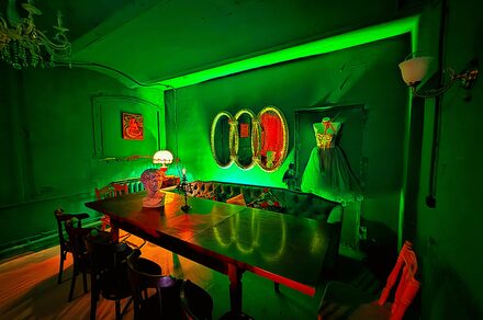 Лофт «Рич» (RICH). Зеленый интерьер с длинным деревянным столом, окруженным черными стульями. На столе лампа с абажуром. На стене три зеркала овальной формы. Угловая софа с подушками. Платье на манекене. Зеленая подсветка. Потолочный светильник.