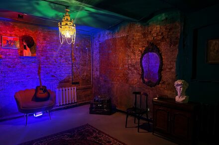 Лофт «Рич» (RICH). Интерьер с кирпичными стенами; потолочное освещение в виде люстры; мягкое кресло с гитарой; зеркало в резной раме; статуя на тумбе; ковёр на полу; разноцветная подсветка; картины на стене; табурет и радиатор вдоль стены; тёмные и теплые цвета.