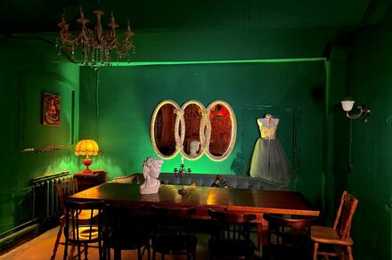 Лофт «Рич» (RICH). Зал с зелеными стенами, длинный деревянный стол с стульями, бюст на столе. Слева торшер с желтым абажуром, на стене картина. Справа платье на манекене. Тройное овальное зеркало на стене. Подвесная люстра.