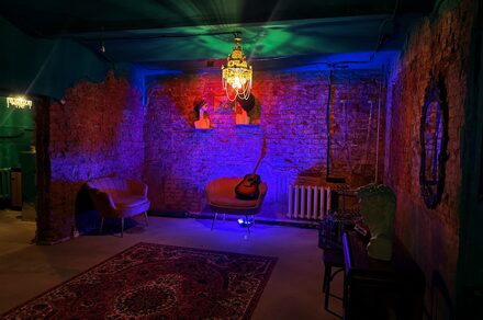 Лофт «Рич» (RICH). Кирпичные стены, два кожаных кресла, кресло с гитарой, потолочная люстра, разноцветное освещение, радиатор, ковер с узором, зеркало на стене, бюст на столике, бетонный пол.