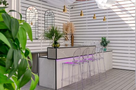 SOK SPACE (Сок Пространство). Лофт МОЙ ПЕРВЫЙ. Барная зона с белыми деревянными панелями и светлыми полами, барная стойка с серой столешницей, прозрачные барные стулья с фиолетовой подсветкой. Подвесные золотистые светильники, зеркала на стене, декор из растений и сухоцветов.