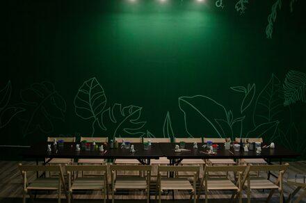 Хендмейдщики. Лофт в лесной тематике. Большой стол с рядами светлых деревянных стульев, расположенный перед зеленой стеной с изображениями листьев. На столе расставлены различные предметы. Светлый деревянный пол, точечное освещение сверху.