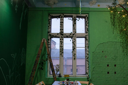 Хендмейдщики. Лофт в лесной тематике. Комната с зелеными стенами, декорированными рисунками растений, и большим окном с занавеской из искусственных листьев. У окна стол с крафтовыми материалами. Справа кирпичная стена, частично оштукатуренная. Деревянная лестница в углу.