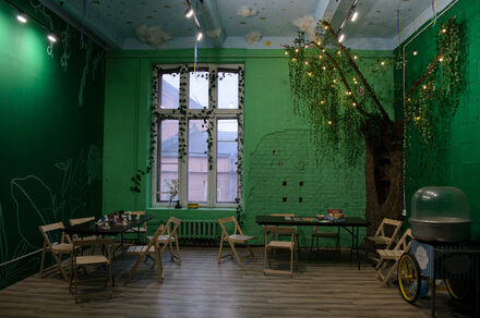 Хендмейдщики. Лофт в лесной тематике. Зеленое помещение с высоким потолком и декоративными элементами. Стены оформлены в виде деревьев с гирляндами. Несколько складных столов и деревянных стульев. Окно с белыми рамами и решеткой. На полу деревянные панели. Освещение точечными светильниками.