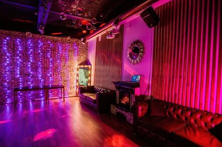 LoftForParty (ЛофтФорПати). Бэкстейдж лофт . Кирпичная стена с фиолетовой подсветкой, пластиковые стулья, диваны из тёмной кожи, деревянный пол, подвесной светильник, зеркало с подсветкой, декоративное зеркало на стене, мебель и стены в теплых тёмных тонах.