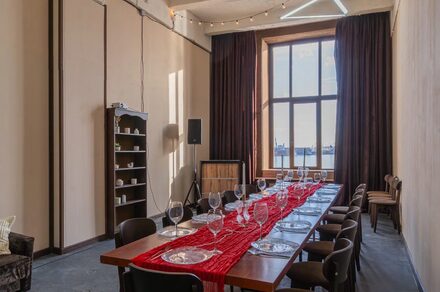 Chocolate Loft(Шоколад Лофт). Зал с видом на Финский залив. Просторное помещение с длинным столом и стульями. На столе красная дорожка, стеклянные бокалы и тарелки. Большое окно с тёмными занавесками. На стенах полки с посудой, акустическая колонка и проектор. Серые полы, светлые стены, подвесное освещение.