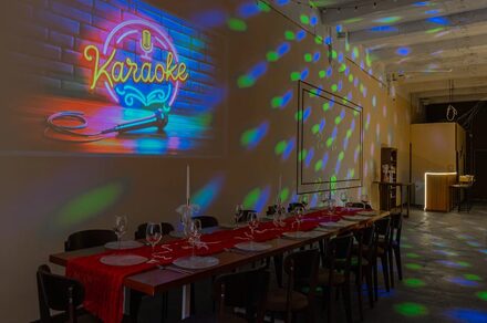 Chocolate Loft(Шоколад Лофт). Зал с видом на Финский залив. Пространство с длинным столом, покрытым красной дорожкой, сервировано бокалами и приборами. На кирпичной стене вывеска "Karaoke" с неоновыми микрофоном. Потолок высокий, с диско-освещением. Справа стойка бара, помещение в темных тонах.