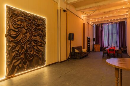 Chocolate Loft(Шоколад Лофт). Зал с видом на Финский залив. Пространство с высокими потолками, длинный стол с красными стульями, диван, деревянный круглый стол. Темные шторы, скульптурное панно с подсветкой, гирлянды на потолке. Светлые стены, темный пол, колонны. Освещение теплое, атмосферное.