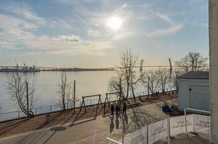 Chocolate Loft(Шоколад Лофт). Зал с видом на Финский залив. Пространство на набережной с видом на водоём и мост. Пешеходная дорожка с деревьями и уличным освещением. Светлое время суток, солнечно. Люди гуляют. Вдали видны портовые краны. Слева ограждение со знаками.