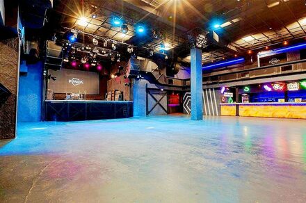 PRAVDA(Правда). Просторный зал на 800 гостей. Широкое внутреннее пространство с бетонным полом. Площадка с оборудованием для выступлений. Стойка бара с подсветкой. Освещение разноцветное, включая прожекторы и неон. Высокий потолок с открытыми металлическими конструкциями. Тёмные стены.