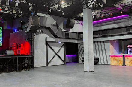 PRAVDA(Правда). Просторный зал на 800 гостей. Просторное помещение с бетонным полом и колонной в центре. Серые стены с черными элементами. Площадка с оборудованием и красной подсветкой. Барная стойка с фиолетовой и розовой подсветкой. Потолочные светильники и аудиосистема.