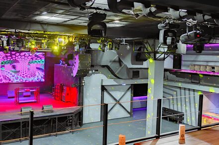 PRAVDA(Правда). Просторный зал на 800 гостей. Клубное помещение с большой сценой, оснащённой экранами и звуковым оборудованием. Верхний этаж с балюстрадой и зонами для сидения. Освещение разноцветное, включая прожекторы. Стены серые, полы тёмные. Потолок в чёрных конструкциях. Отсутствие людей.