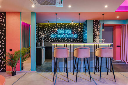 Лофт Шекспир. Стильный дизайнерский зал. Барная зона с высокими стульями и барной стойкой, подсвеченной неоновым текстом «TO BE or not to be». Стены с черно-белым узором, потолочное освещение, яркие цвета, элементы розового и голубого, зеркало, растения в углу.