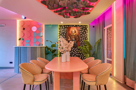Лофт Шекспир. Стильный дизайнерский зал. Розовый овальный стол с бежевыми стульями, на стене портрет в рамке, барная стойка с круглыми элементами, розовый и голубой декор, растения, многоцветное освещение, шторы, потолочный элемент с волнениями.