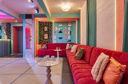 Лофт Шекспир. Стильный дизайнерский зал. Пространство с длинным красным диваном с подушками, круглый столик с бокалами, яркие стены с геометрическими узорами, различные картины, декоративное освещение, разноцветные стены и потолок, плиточный пол светлых оттенков.