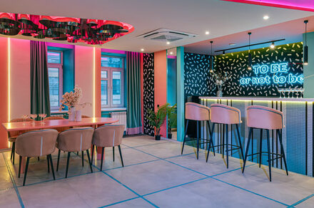 Лофт Шекспир. Стильный дизайнерский зал. Современное праздничное пространство с барной стойкой и столовой зоной. Бар с высокими стульями, неоновая надпись, обеденный стол с мягкими стульями. Яркое декоративное освещение, разноцветные стены, большие окна с шторами, растения в горшках.