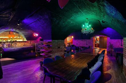 GB Rooms(ГБ Румс). Лофт №1. Кирпичное подвальное помещение с арочным потолком. Длинный деревянный стол, окружённый стульями. Барная стойка на заднем плане. Зелёная и фиолетовая подсветка. Классическая люстра. Пол деревянный. Мягкая мебель вдоль стен.
