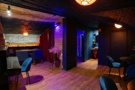 GB Rooms(ГБ Румс). Лофт №1. Подвальное помещение с кирпичными стенами и деревянным полом. Зоны с мягкими синими креслами и диванами, столики. Яркое цветное освещение, красные шторы. Потолок темного цвета. Круглые лампы с теплым светом.