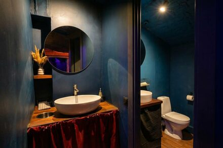 GB Rooms(ГБ Румс). Лофт №1. Ванная комната с синими стенами, круглым зеркалом над раковиной, круглой раковиной на деревянной столешнице, унитазом. Металлический дозатор мыла, красная занавеска под столешницей. Полки с декором, верхнее точечное освещение.
