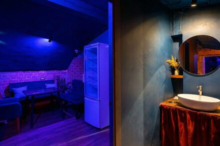 GB Rooms(ГБ Румс). Лофт №1. Комната с синим и красным освещением, кирпичные стены, черная мебель, обеденный стол, диваны, холодильник, круглая раковина с зеркалом, деревянный и керамический декор, занавеска красного цвета, темный деревянный пол.