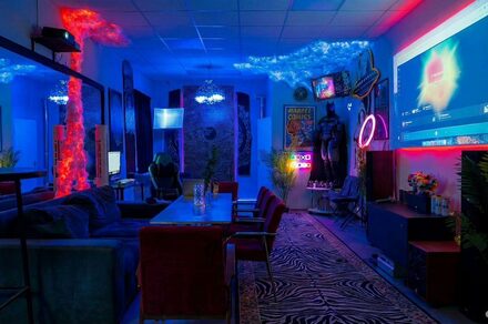 GB Rooms(ГБ Румс). Лофт №2. Квартира с затемнённым освещением, столом и креслами, диванами, неоновыми вывесками и декоративными элементами. На стенах постеры, есть крупный экран для проекций. Используются яркие синие и красные цветовые акценты. На полу ковер с узором.