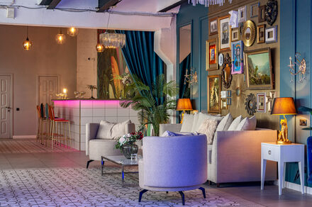Лофт Май. Стильное пространство с верандой. Гостиная с баром в современном стиле. Белые диваны и кресла, стол с цветами. На стене множество картин и зеркал в рамах. Бар с высокими стульями и розовой подсветкой. Потолочные лампы, декоративные растения, синие шторы. Стены бирюзовые, полы плиточные.