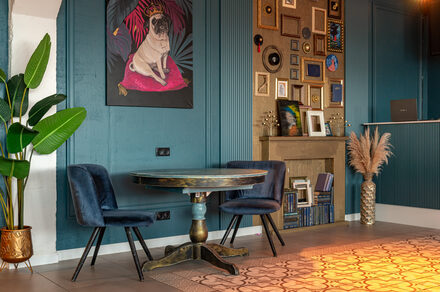 Лофт Май. Стильное пространство с верандой. Угловое пространство с деревянным круглым столом и двумя синими стульями. На стене синие панели, висят картины и фотографии. Полка с декором и несколькими книжными полками. На полу плитка с узором, рядом растение в горшке.