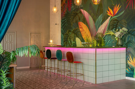 Лофт Май. Стильное пространство с верандой. Барная стойка с белыми квадратными плитками, подсветка розового цвета. Три барных стула с разноцветными спинками. Стена с ярким тропическим рисунком. Зона декорирована зелеными растениями, висят круглые подвесные светильники.