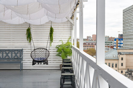 Лофт Май. Стильное пространство с верандой. Терраса с белыми деревянными стенами и крышей, пол серый. Плетеное подвесное кресло, рядом скамейка и табуреты. Растения в горшках. Перила с крестовинами. На заднем плане вид на городскую архитектуру.