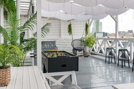 Лофт Май. Стильное пространство с верандой. Открытая терраса с серым деревянным полом, белыми стенами и тканевым потолком. Мебель: диваны, стулья, столики, футбольный стол. Растения в горшках. Вид на город.