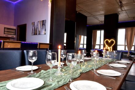 Элегантный лофт с хорошим звуком и балконом. Столовая зона с длинным деревянным столом, сервированным стеклянными бокалами и тарелками. В центре стола зеленая ткань и свечи. Мягкие стулья вдоль стола. На стене картина, сбоку кухня. Светодиодное освещение фиолетового цвета.