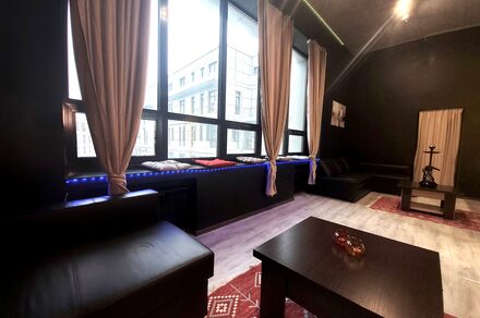 Элегантный лофт с хорошим звуком и балконом. Лофт с чёрными стенами и большими окнами, с бежевыми шторами. Низкие чёрные кожаные диваны и деревянные столы. Красные ковры на светлом полу. На одном из столов расположен кальян. Искусственное освещение, вид на город.